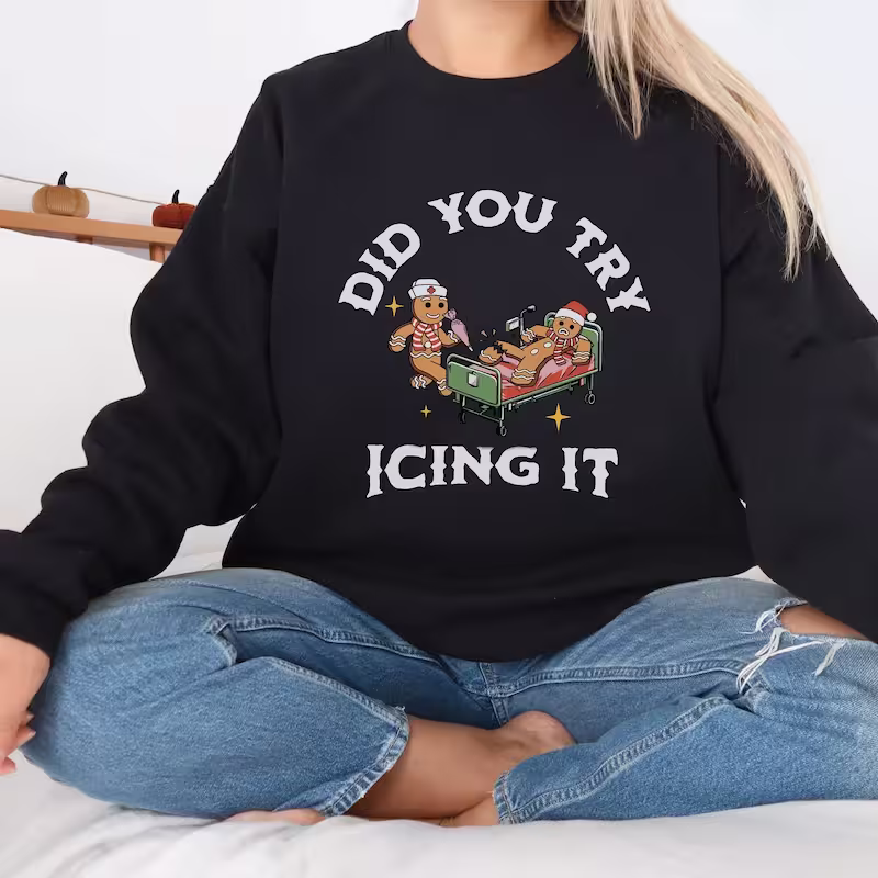 Blonde woman sitting criss-cross in front of holiday decor, wearing the black version of the "Did You Try Icing It?" Funny Christmas Sweatshirt.